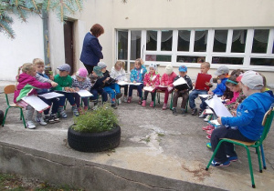 Zdjęcie przedstawia taras przedszkolny, na którym siedzą dzieci w półkolu i rysują zioła rosnące przed nimi.